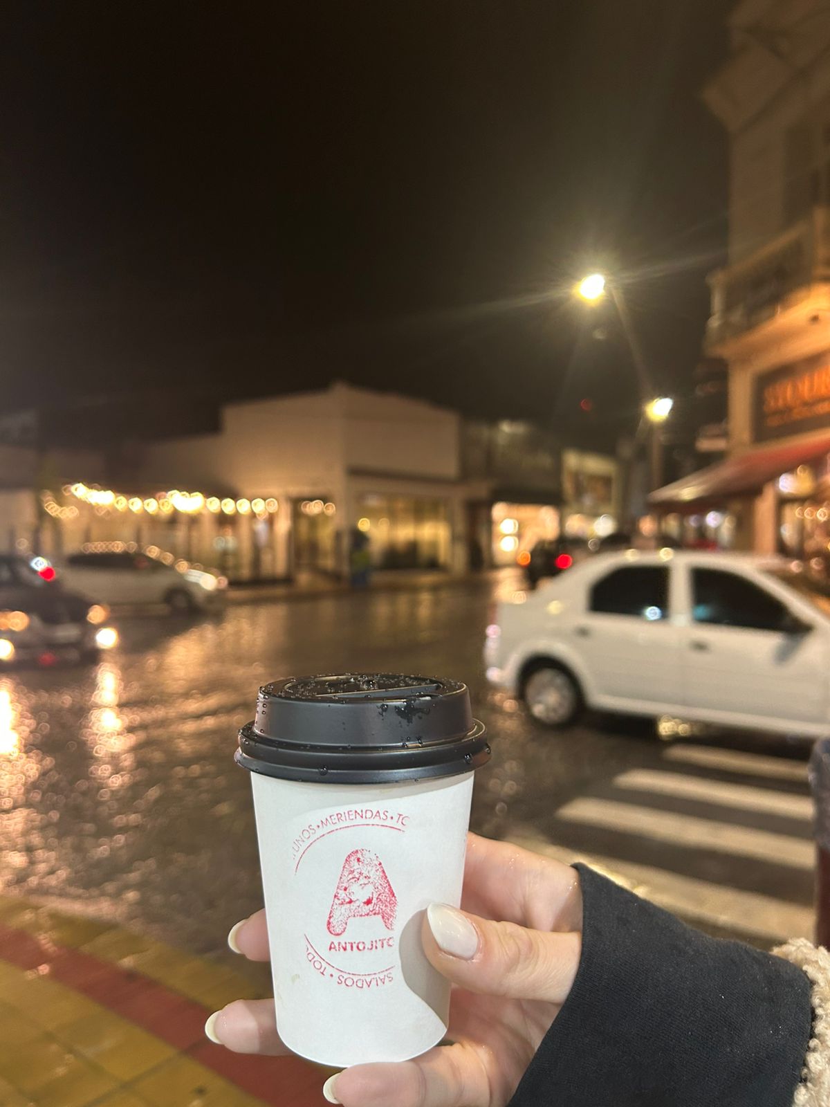 Vaso de café para llevar con el sello de Antojitos, en una calle de Tandil de noche